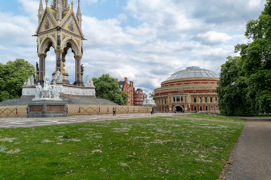 A wide view of a park area featuring a prominent ornate monument with intricate decorative elements, including statues and a gold-colored clock or ornament at the top, situated on a circular base with carved statues and a surrounding low metal fence. To the right, there is a large, round, red-brick building with a domed roof and arched windows, set against a backdrop of green trees and a partly cloudy sky. The foreground shows a grassy lawn with scattered small flowers, bordered by a paved pathway on the right. The scene includes a few pedestrians walking across the open space, with the environment indicating a historical or cultural site, possibly near a city centre. This outdoor setting is adjacent to areas where private waste collection or rubbish removal services such as those provided by [COMPANY_NAME] might assist with site clearance or rubbish removal within public and private spaces, contributing to maintaining the cleanliness and preservation of such public parks and historic landmarks.