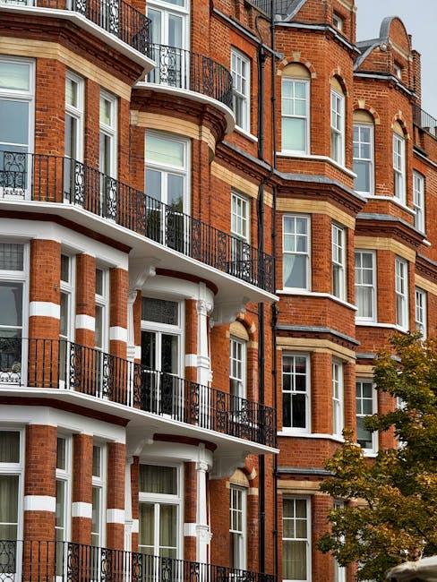 The image depicts a multi-storey red brick residential building with white and beige decorative accents around the windows and balconies. The building features several protruding bay windows with white frames, and the balconies have black wrought iron railings. The facade includes curved architectural elements and a combination of brickwork patterns, creating a classic London-style appearance. In the foreground, there is a tree with green leaves partially covering the lower part of the building, and the setting appears to be an urban residential area with natural daylight illuminating the scene. The overall look of the building reflects traditional brick construction with modern detailing, typical of city centre residences that may benefit from private waste collection or external rubbish removal services, as offered by companies like rubbishremovalwestkensington.co.uk, to manage waste efficiently alongside their urban environment.