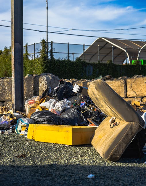 A daytime outdoor scene shows an accumulated pile of rubbish scattered across a gravel surface, consisting of black plastic trash bags, loose paper, and miscellaneous debris. The bags are partially torn open, revealing waste inside, and some items are spilling out onto the ground. Prominent in the foreground is a large, beige, weathered car seat or cushion leaning against the trash, next to a yellow plastic container lying on its side. Behind the rubbish, there is a low stone wall made of irregularly shaped bricks or stones, partly obscuring a fence and some greenery. To the left, a metal post and overhead power lines are visible against a blue sky with scattered clouds. In the background, a sports or tennis court with a curved, transparent cover and supporting structures can be seen, indicating a suburban or urban outdoor environment. The scene conveys an example of unauthorized waste accumulation set against an outdoor sports facility, highlighting the need for effective rubbish removal services such as those offered by [COMPANY_NAME].