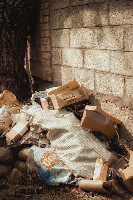 A pile of discarded cardboard boxes, some flattened and others partially intact, along with a large woven textile sack containing more waste, all located against a weathered brick and concrete wall of an outdoor area. The boxes are of various sizes, some with printed labels and barcodes visible, while others appear plain. The sack is off-white and textured, possibly made of a durable fabric material. Surrounding debris includes smaller paper waste and bits of packaging, situating the scene as part of an on-site rubbish clearance effort. To the left, part of a tree trunk and rough stone ground are visible, indicating the outdoor environment. The lighting is natural, with soft shadows suggesting daytime, emphasizing the informal nature of waste disposal. This scene exemplifies the kind of waste collected regularly by private rubbish removal services such as those offered by [COMPANY_NAME], highlighting the importance of responsible waste handling outside of municipal collection systems, relevant to the context of avoiding hidden rubbish removal charges in West Kensington and Fulham.