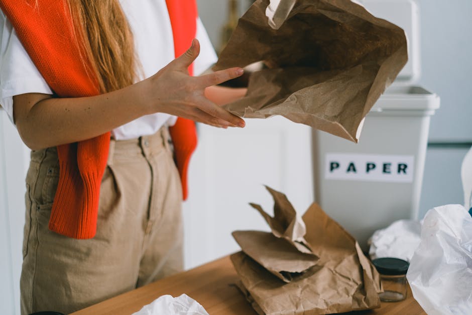 A person wearing a white shirt and beige pants, partially visible, is holding an open brown paper bag with both hands, preparing to dispose of waste. The individual has a red knitted cardigan draped over their shoulders. On a wooden table in front, there are additional crumpled brown paper bags and white plastic waste, with a background featuring a white recycling bin labeled 'PAPER' in black letters. The environment appears to be an indoor office or kitchen space, illuminated by natural light, with a neutral, tidy setting. The scene suggests activities related to waste sorting or on-site rubbish disposal, consistent with independent rubbish collection or recycling activities managed outside of municipal waste systems, possibly supported by private waste management services such as those found at rubbishremovalwestkensington.co.uk.