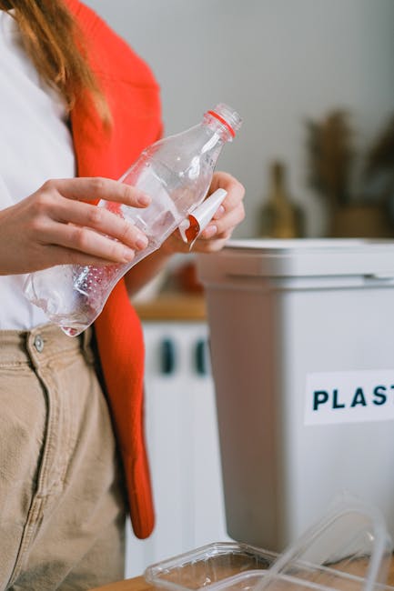 Close-up image of a person wearing a red cardigan and beige trousers, holding a transparent plastic bottle with a white cap, positioning it over a white recycling bin labeled 'PLASTIC'. The background shows an indoor setting with a washing machine or dryer, and some blurred objects indicating a domestic environment. The scene reflects the act of preparing plastic waste for recycling, aligning with environmentally-conscious rubbish removal practices, potentially managed by services like those offered by [COMPANY_NAME]. The focus on the plastic bottle, its texture, and transparent material highlights the emphasis on recycling and waste separation, useful in discussions about private waste handling and on-site rubbish collection solutions within residential spaces.