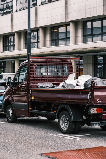 A small, dark red pickup truck with an open cargo bed parked on a city street in front of a modern office building with large glass windows. The cargo bed is loaded with various dumped waste materials, including black rubbish bags, white plastic bags, and loose debris. The truck features a metal mesh barrier at the front of the cargo bed, providing protection and separation from the cab area. The vehicle’s body shows a smooth, glossy finish with some signs of use, and metallic wheels with visible hubcaps. Surrounding the truck, the environment includes other parked vehicles, such as part of a white van in the background, and the street is paved with asphalt. A streetlamp pole is positioned near the truck, partially obscuring some of the building’s façade. This scene reflects a scenario of private waste disposal, likely associated with rubbish removal services like those offered by companies such as rubbishremovalwestkensington.co.uk, emphasizing on-site clearance and alternative waste handling methods within an urban context.