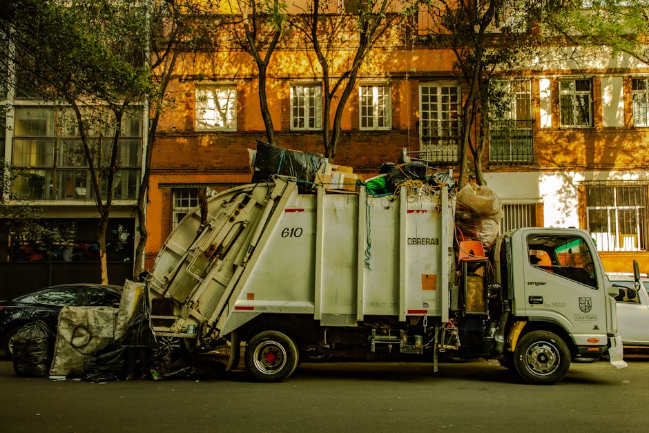 A side view of a white rubbish collection truck parked on a city street during daytime, with various waste materials loaded into its open-top container. The truck's body features a hydraulic lift system and is marked with the number 610, while the cab is fitted with a small logo and identification details. The waste load includes black garbage bags, cardboard boxes, plastic containers, and assorted debris. The scene is set in front of a residential building with a brick façade, multiple windows, and some green foliage from nearby trees casting shadows across the building and street. The environment appears calm with natural daylight illuminating the scene, reflecting typical private waste collection or rubbish removal activities in an urban setting and supporting the context of independent rubbish disposal services.