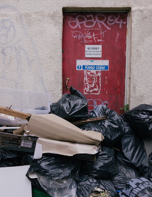 Piles of black rubbish bags and discarded cardboard boxes stacked against a weathered external wall beneath a small, rectangular window opening. The bags appear to be filled with household or rubbish waste and are situated on a pavement or alleyway outside a building. The background features a red metal door covered in graffiti tags and stickers, including a white sign signifying 'No Money' and a blue and white sign that reads 'Keep clear.' The scene suggests a private rubbish collection or on-site clearance activity, with the waste awaiting removal by waste management services such as those offered by rubbishremovalwestkensington.co.uk. The environment appears unkempt, with the waste covering most of the lower section of the wall, indicating an outdoor disposal situation typical of independent waste handling outside commercial or residential premises.