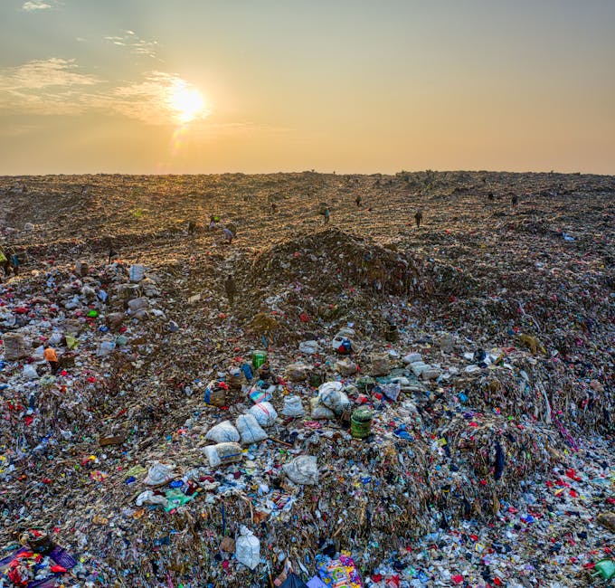A vast landfill site under a setting sun, displaying large mounds of mixed waste materials including plastic bottles with green caps, paper, and other debris, spread across a barren, uneven terrain. The waste appears densely packed, creating towering piles with visible textures of crinkled plastic, rigid cardboard, and soil. Scattered small objects and fragments are noticeable throughout the site, with some waste spilling over the edges of the mounds. In the background, a few distant figures can be seen walking across the site, contributing to the impression of a large-scale waste management operation typical of private rubbish collection or alternative disposal methods. The sky is illuminated by a warm, golden glow from the low sun, casting long shadows and emphasizing the expansive, unmanaged landscape, which visually contrasts with the controlled waste removal services offered by companies like rubbishremovalwestkensington.co.uk. The scene highlights the environmental impact of waste accumulation and underscores the importance of proper rubbish clearance in urban environments.