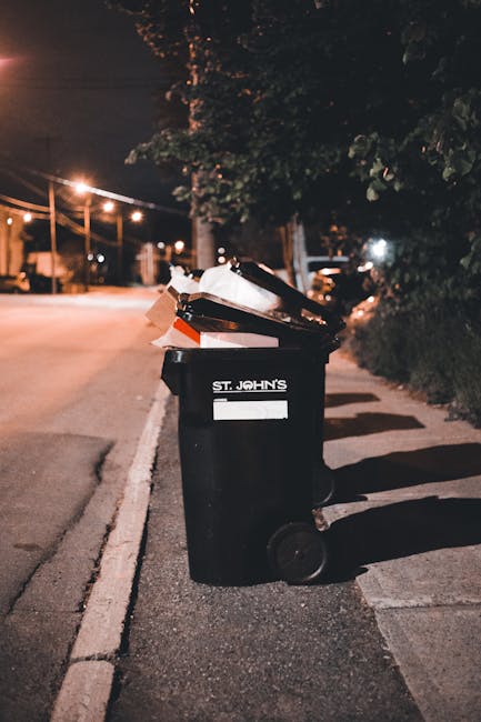 A black wheeled rubbish bin labeled 'St. John's' is positioned on a city sidewalk near the curb, filled with various discarded items, including paper, cardboard, and plastic materials, some of which are overflowing and leaning outward. The bin is situated on a paved pathway adjacent to a street illuminated by warm streetlights, casting soft shadows on the ground. Behind the bin, a row of trees with leafy branches partially obscures the background, which includes parked cars and faint distant lighting, suggesting an evening or nighttime setting. The scene reflects an environment where private waste collection services may handle refuse disposal outside of municipal systems, emphasizing the need for proper waste management and rubbish removal practices. The overall atmosphere is quiet and urban, with the focus on the bin’s contents and its placement in a typical residential or mixed-use area.