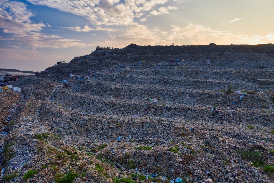 A large landfill site with an expansive, sloped terrain filled with layers of mixed waste and rubble, predominantly composed of dark, rough-textured materials such as broken concrete, plastic, and discarded debris. The waste is spread unevenly across the hillside, with some areas showing more tightly packed rubbish, while others have scattered patches of green vegetation growing through small gaps. In the background, a few small structures and piles of waste can be seen at the top of the mound, indicating active waste deposition or monitoring stations. The sky above is partly cloudy with a soft, warm glow from the setting or rising sun, casting diffuse light over the scene, highlighting the rugged, uneven surface of the landfill. This image visually reflects the scale and environmental impact of unmanaged waste accumulation, aligning with discussions on alternative waste handling and rubbish disposal challenges, as often considered in private rubbish removal services. Natural environmental context of ground and sky are evident, emphasizing the substantial size and layered composition of the landfill, typical of large-scale waste management operations.