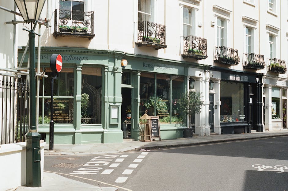 The image shows a street scene in an urban area featuring a row of commercial shops with large glass display windows. The storefront on the left is painted in a pastel green color, with decorative molding and a sign indicating a flower shop. In front of this shop, there are potted plants and a small blackboard sign, typical of a boutique shop front. Adjacent to it, a black-painted shop has a sleek, modern appearance with large windows and minimal decoration. The buildings are multi-story with white facades and evenly spaced windows, each with small, wrought-iron balconies adorned with flower pots. A lamppost stands on the sidewalk near the green shop, and a street sign with a red and white no-entry barricade is visible near the corner. The street has bicycle lane markings and pedestrian crossings, and the scene is illuminated by natural daylight, creating a clean, tidy appearance consistent with well-maintained urban commercial areas. The context suggests an environment where alternative waste handling, such as private rubbish removal services by companies like rubbishremovalwestkensington.co.uk, might be relevant for shop and office refuse collections.