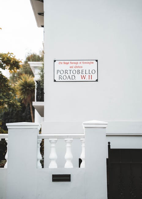 A white exterior wall of a residential building featuring a decorative white balustrade with three evenly spaced, turned balusters positioned in front of a black gate. Mounted on the wall is a rectangular street sign with a black border and red lettering indicating 'Portobello Road, W11,' and additional text referencing the Royal Borough of Kensington and Chelsea. The background shows some elements of greenery, including trees and shrubs, which contrast against the light-colored wall. The scene is captured with soft, natural lighting, emphasizing the clean, smooth surface of the wall and the polished finish of the balustrade. This setting aligns with the context of private property and independent waste management, as often managed by services like rubbish removal specialists operating in West Kensington or similar London districts, emphasizing the urban residential environment where on-site clearance may take place for rubbish or bulky waste collection purposes.
