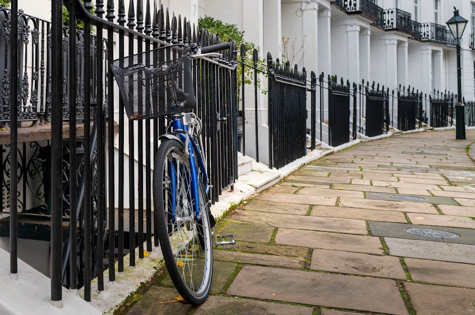 In the foreground, a blue bicycle with a black front basket leans against a black metal fence that features pointed finials along the top. The bicycle’s frame, handlebar, and front wheel are partially obscured by the fence, with the bike situated on a pavement composed of large, irregularly shaped stone slabs showing subtle weathering and moss growth in the joints. Behind the fence, a white building with classical architectural elements such as columns and decorative railings on the balcony can be seen, with a small green shrub peeking over the fence to the left. The scene is evenly lit with natural daylight, emphasizing the textures of the stone pavement, the wrought iron fence, and the painted surface of the bicycle. This setting reflects a typical residential street scene in West Kensington, where private waste management or rubbish removal services such as those offered by rubbishremovalwestkensington.co.uk could be relevant for maintaining clear pathways and frontages for local homes, especially during rubbish collection or clearance activities taking place in similar urban environments.